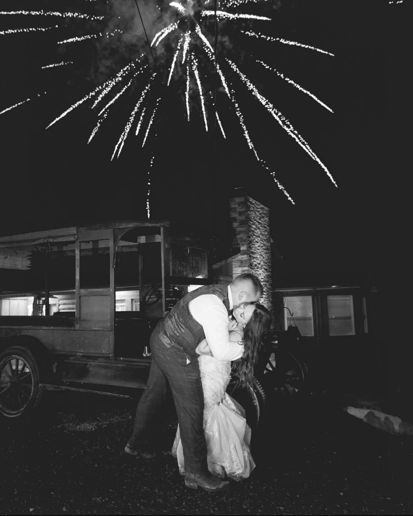 Newlyweds kissing beside a vintage car under fireworks